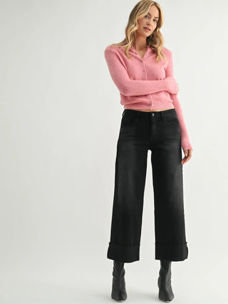A woman modeling a pink ribbed knit cardigan with black wide-leg cropped jeans and black ankle boots, standing against a light gray studio background.