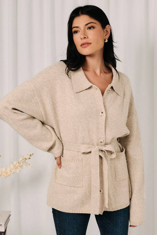 A woman wearing a light beige knit button-front cardigan with a tie waist belt and front patch pockets, paired with dark blue jeans, set against a soft white curtain background.