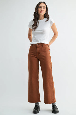 A woman modeling a pair of rust-brown wide-leg cropped jeans with a white t-shirt and black loafers, standing against a light gray studio background.