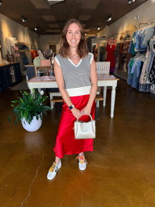 A woman stands in a clothing boutique wearing a sleeveless black and white striped top, a bright red satin midi skirt, and white sneakers with colorful laces, holding a small white handbag.