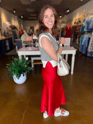 A woman posing inside a boutique shop, wearing a black and white striped sleeveless top with a white bow detail, a vibrant red satin skirt, and white sneakers, while carrying a cream-colored handbag.