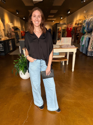 A woman stands in a clothing boutique wearing a black wrap style, faux tucked  blouse, light-wash wide-leg jeans, and black shoes, holding a black woven clutch.
