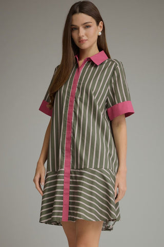 A woman modeling a short-sleeve collared shirt dress with vertical olive green and white stripes and contrasting pink trim on the collar, placket, and cuffs, against a plain gray studio background.