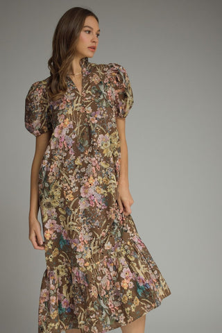 A woman modeling an eyelet brown midi dress with a dense multicolor floral print, featuring puff sleeves and a ruffled hem, set against a plain gray studio background.