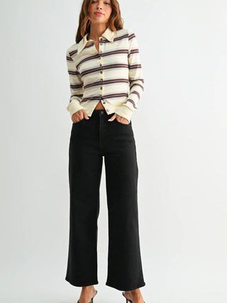 A woman modeling a cream-colored knit striped button-up shirt with brown and dark stripes, paired with black wide-leg cropped pants, against a plain light gray studio background.