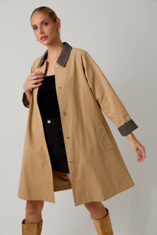 A woman models a mid-length tan trench coat with a contrasting dark checkered collar and cuffs, worn over a black dress and tan boots against a light gray studio background.