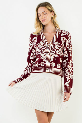 A woman modeling a burgundy and cream patterned knit cardigan with a V-neck and striped trim, paired with a white pleated skirt, against a plain white studio background.